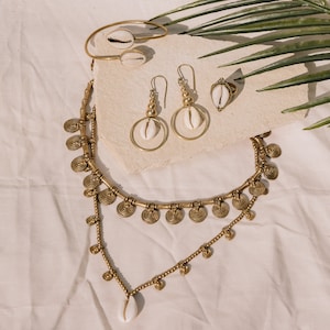 Puede incluir: Una colecci&oacute;n de joyas doradas con conchas de cauri. El conjunto incluye un collar, pendientes, un anillo y una pulsera. El collar tiene un dise&ntilde;o en capas con detalles en forma de moneda. Los pendientes tienen un dise&ntilde;o de aro. Las joyas se exhiben sobre una superficie neutra.