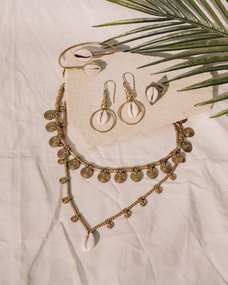 Puede incluir: Una colecci&oacute;n de joyas doradas con conchas de cauri. El conjunto incluye un collar, pendientes, un anillo y una pulsera. El collar tiene un dise&ntilde;o en capas con detalles en forma de moneda. Los pendientes tienen un dise&ntilde;o de aro. Las joyas se exhiben sobre una superficie neutra.