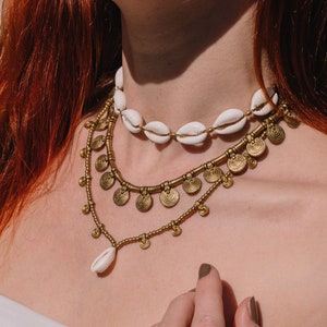 Può includere: Una collana a strati con un choker di conchiglie bianche, una catena dorata con piccoli ciondoli rotondi e una catena dorata delicata con un solo ciondolo di conchiglia bianca.