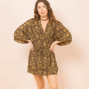 Op de afbeelding: Zwart en goud bloemenprint kimono-stijl jurk met een V-hals en strik in de taille. De jurk heeft wijde mouwen en een mini-jurk lengte. Het bloemenpatroon is goudkleurig op een zwarte achtergrond. Het model draagt een zwarte choker.