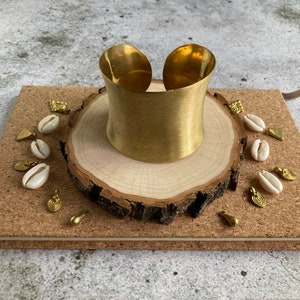 K&ouml;nnte beinhalten: Ein goldfarbenes Metall-Armband mit glatter, geb&uuml;rsteter Oberfl&auml;che. Das Armband wird auf einer Holzscheibe mit wei&szlig;en Muscheln und goldenen Akzenten pr&auml;sentiert.