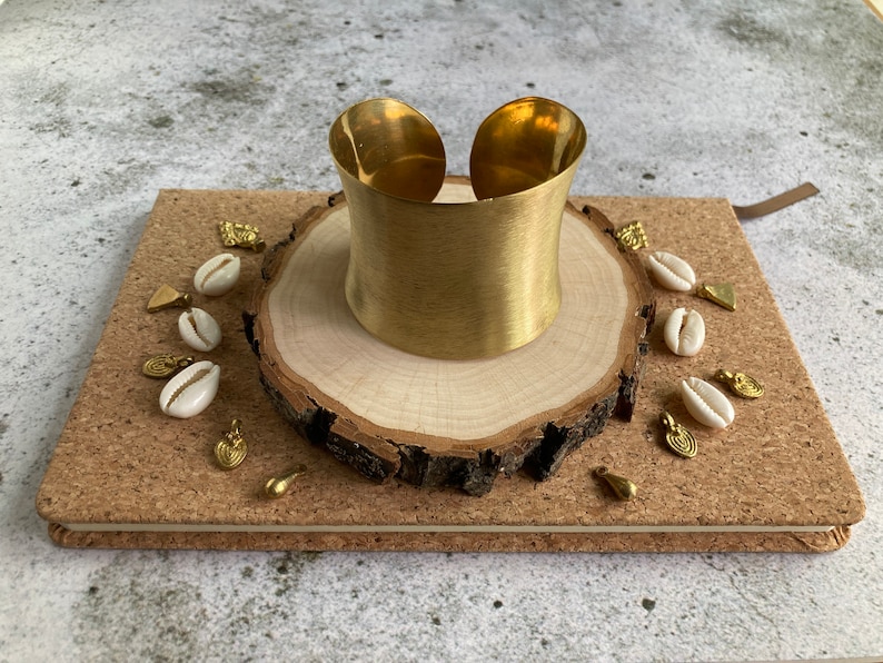 K&ouml;nnte beinhalten: Ein goldfarbenes Metall-Armband mit glatter, geb&uuml;rsteter Oberfl&auml;che. Das Armband wird auf einer Holzscheibe mit wei&szlig;en Muscheln und goldenen Akzenten pr&auml;sentiert.