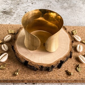 K&ouml;nnte beinhalten: Ein breites goldenes Metall-Armband mit glatter, geb&uuml;rsteter Oberfl&auml;che. Das Armband wird auf einer Holzscheibe mit kleinen goldenen und wei&szlig;en Dekorationen pr&auml;sentiert.