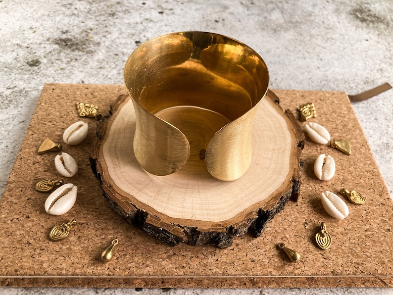 K&ouml;nnte beinhalten: Ein breites goldenes Metall-Armband mit glatter, geb&uuml;rsteter Oberfl&auml;che. Das Armband wird auf einer Holzscheibe mit kleinen goldenen und wei&szlig;en Dekorationen pr&auml;sentiert.