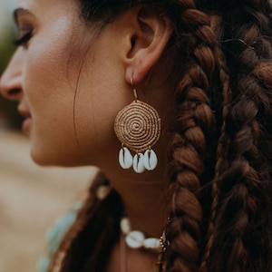 Könnte beinhalten: Nahaufnahme einer Person, die goldfarbene Ohrringe trägt. Die Ohrringe haben ein Spiraldesign mit drei herabhängenden weißen Muscheln. Die Person hat lange, geflochtene braune Haare und trägt ein blau-grünes Oberteil. Die Ohrringe sind im Bohème-Stil.