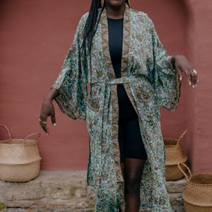 Könnte beinhalten: Ein langer, fließender Kimono in Türkis-, Braun- und Lilatönen mit floralem Muster. Der Kimono hat weite Ärmel und einen passenden Gürtel. Die Person trägt ein schwarzes Outfit. Weidenkörbe sind im Hintergrund.