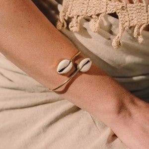 Puede incluir: Un brazalete dorado con dos conchas de cauri blancas y beige. El brazalete se lleva en un brazo, con un fondo de tonos neutros. Las conchas tienen un aspecto natural y texturizado, a&ntilde;adiendo un toque de estilo costero.