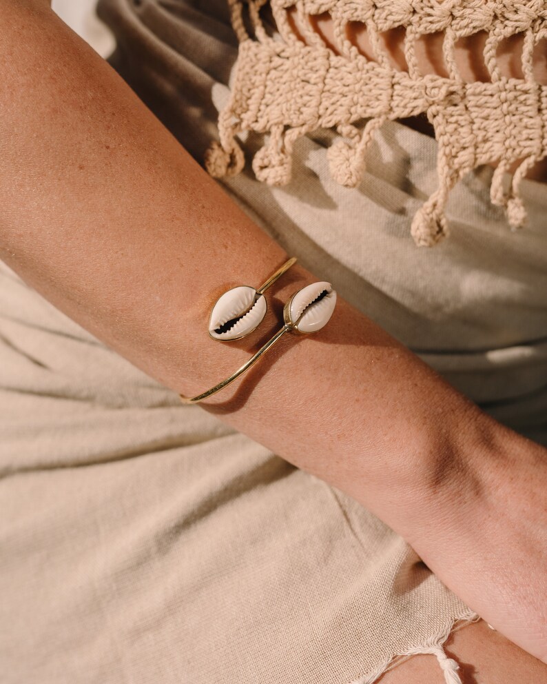 Puede incluir: Un brazalete dorado con dos conchas de cauri blancas y beige. El brazalete se lleva en un brazo, con un fondo de tonos neutros. Las conchas tienen un aspecto natural y texturizado, a&ntilde;adiendo un toque de estilo costero.
