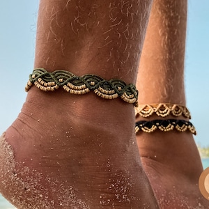 Könnte beinhalten: Zwei Fußkettchen mit grünem und schwarzem Band und goldenen Perlen. Die Fußkettchen befinden sich an den Knöcheln einer Person. Der Hintergrund ist ein Strand mit blauem Wasser und Sand.