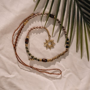 Può includere: Una collana di corda intrecciata marrone e verde con perline di legno e un ciondolo a forma di stella dorata. La collana è appoggiata su uno sfondo di tessuto bianco.