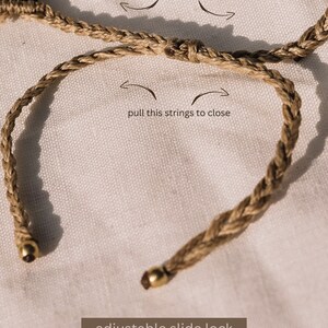 May include: A brown braided cord bracelet with a gold adjustable slide lock. The bracelet is shown on a white fabric background. The text "pull this strings to open" and "pull this strings to close" is shown with arrows indicating how to open and close the bracelet.