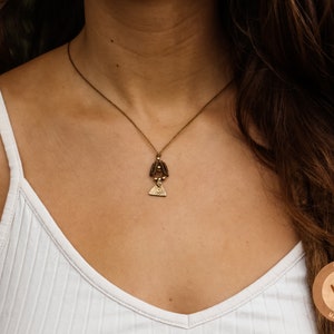 Peut inclure: Une femme portant un débardeur blanc avec un collier en or délicat orné d'un petit pendentif complexe. Le pendentif est un design superposé avec une forme triangulaire en bas et un cercle avec une petite perle en haut. Le collier est de style chaîne simple.