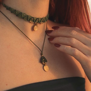 May include: A green macrame choker necklace with a gold spiral pendant and a second gold pendant with a spiral design. The necklace is worn by a person with red hair.