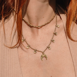 Può includere: Una collana dorata a più fili con un ciondolo a forma di mezzaluna e piccoli ciondoli a forma di moneta. La collana è indossata da una persona con i capelli rossi.