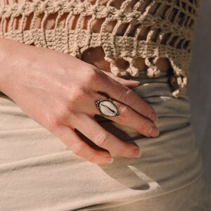 Puede incluir: Un anillo de lat&oacute;n con un centro ovalado de concha blanca. El anillo est&aacute; en una mano, con un top de ganchillo beige en el fondo. El dise&ntilde;o del anillo presenta un borde detallado y texturizado.