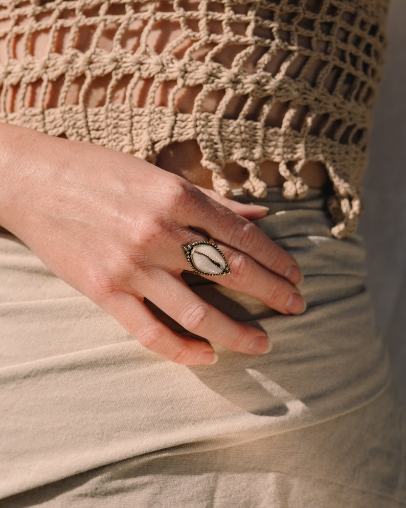 Puede incluir: Un anillo de lat&oacute;n con un centro ovalado de concha blanca. El anillo est&aacute; en una mano, con un top de ganchillo beige en el fondo. El dise&ntilde;o del anillo presenta un borde detallado y texturizado.