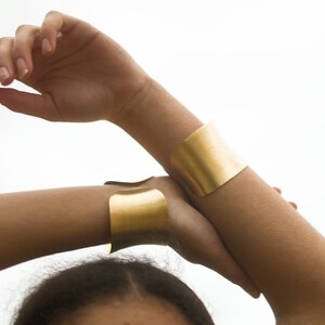 K&ouml;nnte beinhalten: Zwei goldene Metallarmb&auml;nder am Arm einer Person. Die Armb&auml;nder sind breit und flach und beide aus dem gleichen Metall.