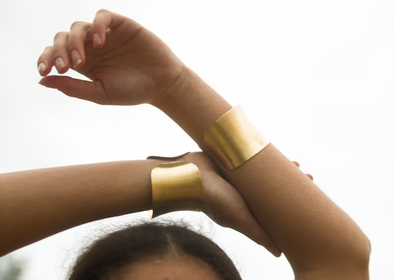 K&ouml;nnte beinhalten: Zwei goldene Metallarmb&auml;nder am Arm einer Person. Die Armb&auml;nder sind breit und flach und beide aus dem gleichen Metall.