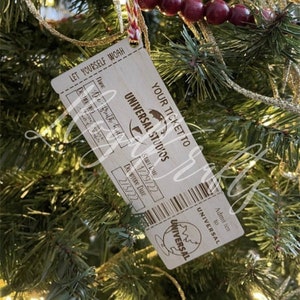 May include: A wooden ornament shaped like a vintage ticket stub for Universal Studios. The ornament is brown with white text and a black and white logo. The text on the ornament reads "Let yourself woah", "Your ticket to Universal Studios", "Start Date 1/29/22", "Date Wine Date 2/13/22", "Admit ten to Universal".