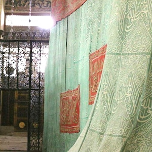 Op de afbeelding: Een interieur met een groene houten muur versierd met rode rechthoekige textiel. Een groen gordijn met patroon en witte kalligrafie hangt rechts. Een decoratief zwart metalen hek is zichtbaar op de achtergrond, met zonlicht.