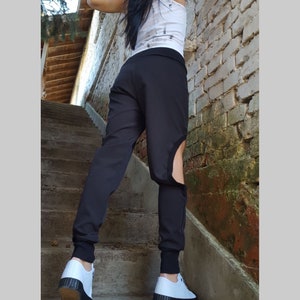 May include: Black trousers featuring a distinctive cutout detail on the lower leg. The trousers have a fitted shape with elasticated cuffs at the ankles. The person is wearing white trainers with black soles. The background includes a brick wall and concrete steps.