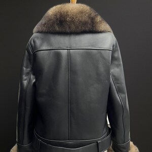 May include: Black leather jacket with a brown fur collar and cuffs. The jacket has a belt at the waist and is displayed on a hanger. The back of the jacket is visible, showcasing the stitching details.