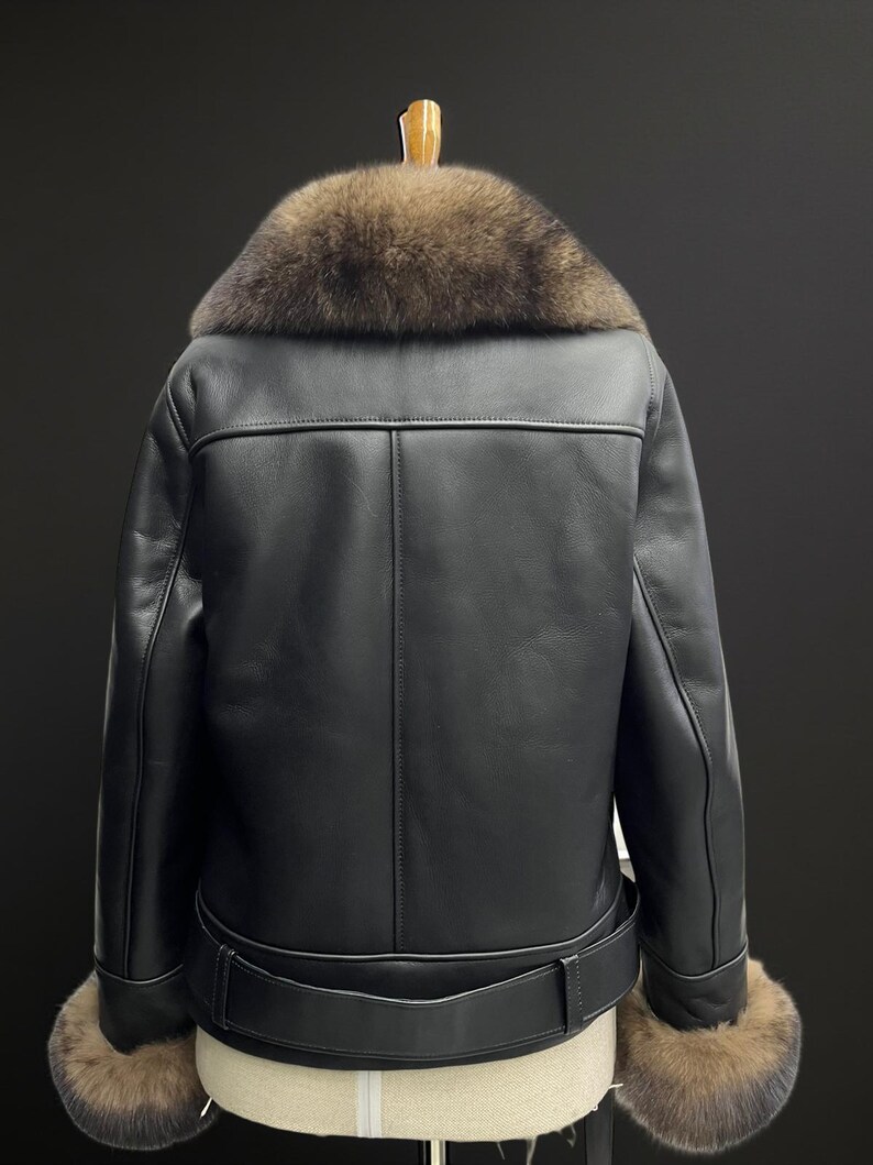 May include: Black leather jacket with a brown fur collar and cuffs. The jacket has a belt at the waist and is displayed on a hanger. The back of the jacket is visible, showcasing the stitching details.