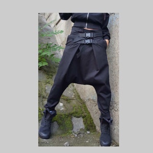 May include: Black harem trousers with a high waist and buckle details. The trousers have a relaxed fit with a drop crotch and are paired with black boots. The model is wearing a black jacket.