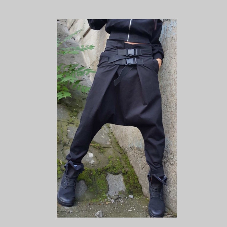 May include: Black harem trousers with a high waist and buckle details. The trousers have a relaxed fit with a drop crotch and are paired with black boots. The model is wearing a black jacket.