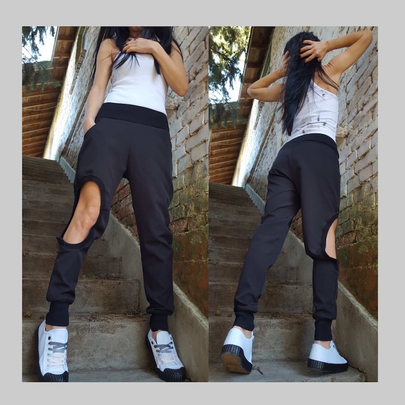 May include: Black trousers with a distinctive leg cutout, worn with a white vest top and white trainers. The trousers feature a broad black waistband and fitted cuffs at the ankles. The model is standing on outdoor steps.
