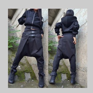 May include: Black hooded jacket with a zip, worn with black harem trousers featuring a belt with buckles. The outfit includes black boots. The clothing is made of a solid black material.
