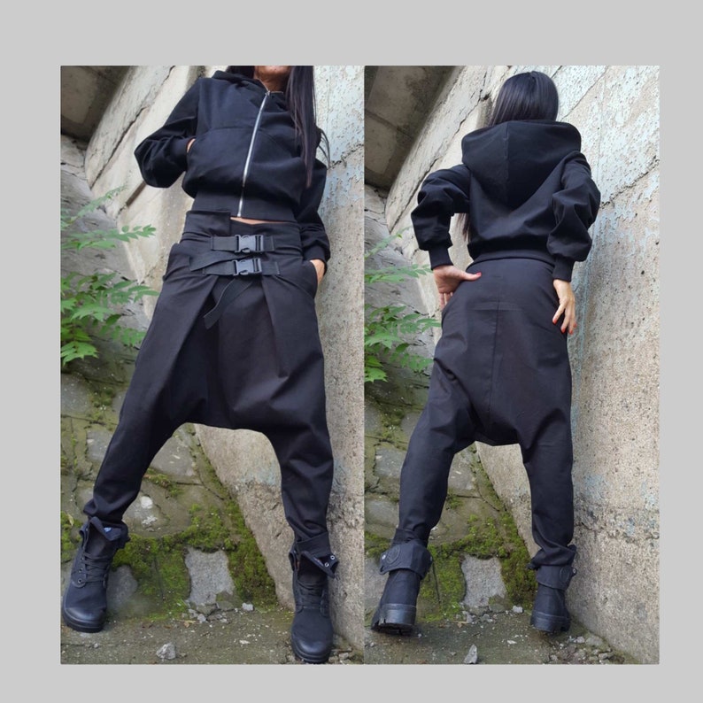 May include: Black hooded jacket with a zip, worn with black harem trousers featuring a belt with buckles. The outfit includes black boots. The clothing is made of a solid black material.