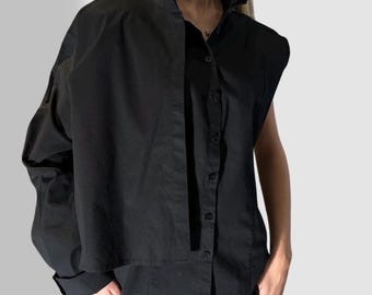 May include: Black button-down shirt with a high, pointed collar and long sleeves. The shirt has a unique design with one sleeve cut off. The shirt is paired with black pants. A small red detail is visible on the shirt.