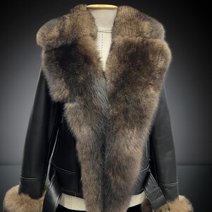 May include: A black leather jacket with a large fur collar and fur cuffs. The jacket has a belt at the waist and is displayed on a mannequin. The fur is a mix of brown and gray tones.