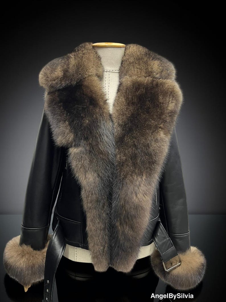 May include: A black leather jacket with a large fur collar and fur cuffs. The jacket has a belt at the waist and is displayed on a mannequin. The fur is a mix of brown and gray tones.