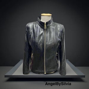 Può includere: Giacca in pelle nera con texture a pelle di serpente, esposta su un manichino. La giacca ha un collo alto, una cerniera a tutta lunghezza e dettagli dorati. Il marchio "AngelBySilvia" è visibile in basso.