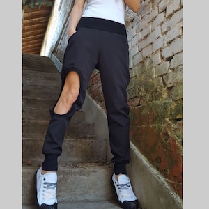 May include: Black joggers with a distinctive cutout detail on the left leg. The trousers feature a broad black waistband and fitted cuffs at the ankles. The person is wearing white trainers with black soles and grey laces. The background includes a brick wall and concrete steps.