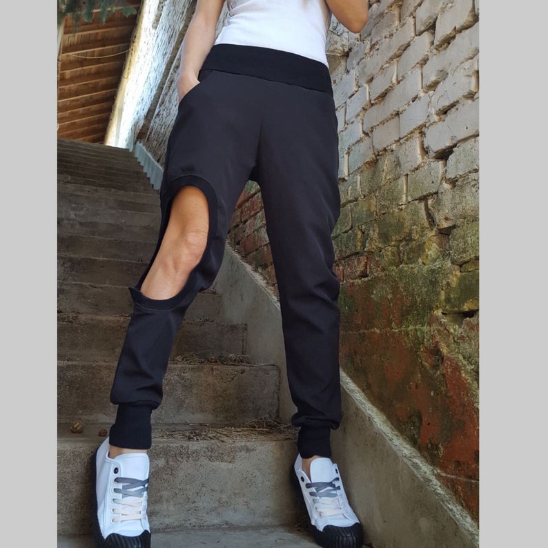 May include: Black joggers with a distinctive cutout detail on the left leg. The trousers feature a broad black waistband and fitted cuffs at the ankles. The person is wearing white trainers with black soles and grey laces. The background includes a brick wall and concrete steps.