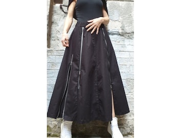 May include: A black maxi skirt with silver zippers and side slits. The skirt has a high waist and is paired with a black top and white sneakers. The outfit is set against a textured gray wall.