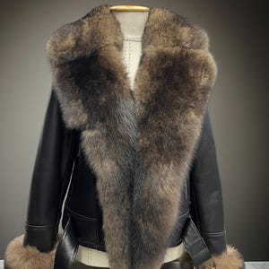 May include: A black leather jacket with a large fur collar and fur cuffs. The jacket has a belt at the waist and a buckle closure. The fur is a mix of brown and gray tones. The jacket is displayed on a mannequin.