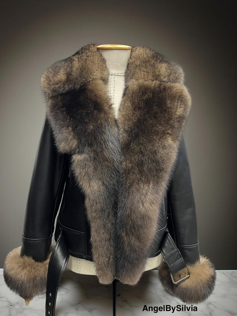 May include: A black leather jacket with a large fur collar and fur cuffs. The jacket has a belt at the waist and a buckle closure. The fur is a mix of brown and gray tones. The jacket is displayed on a mannequin.