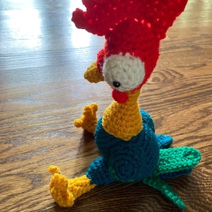 May include: A crocheted rooster toy with red, yellow, blue, and green yarn. The rooster has a large white eye with a black pupil.