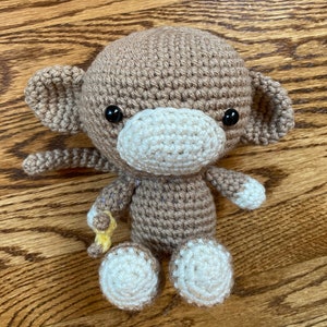 May include: A crocheted monkey toy with brown fur, white face, and black eyes. The monkey is holding a yellow crocheted banana.