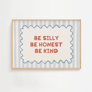 May include: A framed print featuring the words "BE SILLY BE HONEST BE KIND" in red. The text is within a light blue wavy border on a cream background with light blue vertical stripes. The frame is a light wood colour.