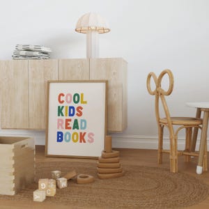May include: A framed print with the text "COOL KIDS READ BOOKS" in colorful letters. The print is hanging on a light wood cabinet with a white lamp on top. There is a brown rug on the floor with wooden toys and a wooden chair.