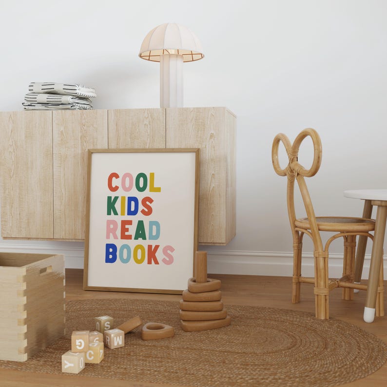 May include: A framed print with the text "COOL KIDS READ BOOKS" in colorful letters. The print is hanging on a light wood cabinet with a white lamp on top. There is a brown rug on the floor with wooden toys and a wooden chair.