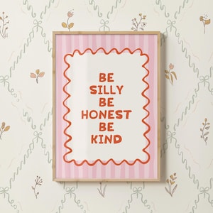 May include: A framed art print with the words "BE SILLY BE HONEST BE KIND" in red on a white background with a wavy red border. The print has a pink striped background and a light brown wooden frame. The background is a cream color with a floral pattern.
