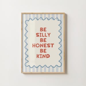 Puede incluir: Una impresi&oacute;n art&iacute;stica enmarcada con un fondo beige claro y un borde a rayas azules y blancas. La impresi&oacute;n presenta las palabras "BE SILLY BE HONEST BE KIND" en letras rojas. El marco es de color marr&oacute;n claro.