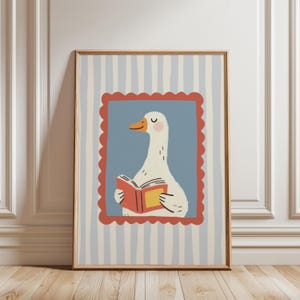 May include: A framed art print featuring a whimsical illustration of a white goose reading a book. The goose is set against a blue background within a red scalloped border, framed by a light wood frame. The background has vertical stripes.