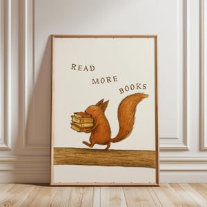 May include: A framed print with a cartoon squirrel carrying books. The squirrel is brown with a bushy tail, walking on a branch. The words "READ MORE BOOKS" are printed above the squirrel. The background is off-white, and the frame is a light brown.